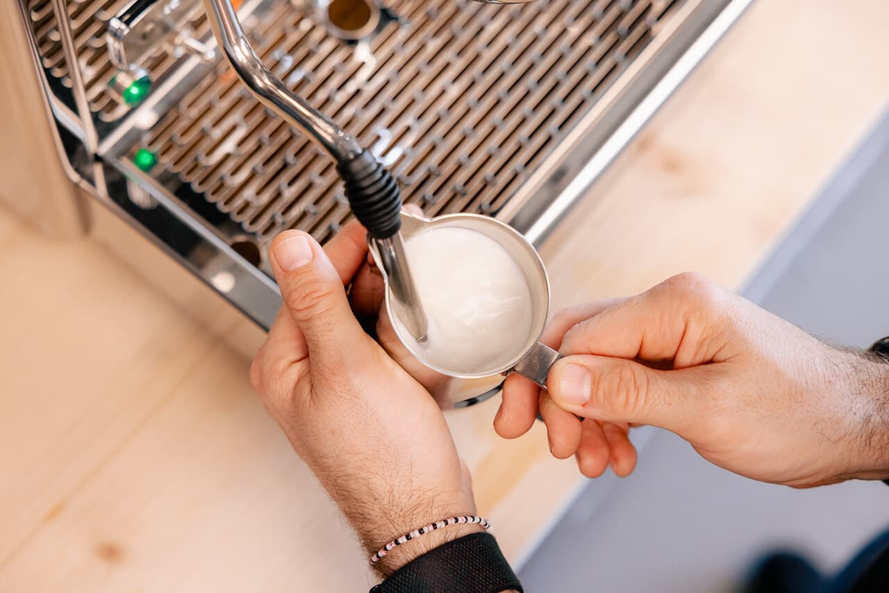 Hände schäumen Milch mit Dampfdüse an Heim-Espressomaschine – Edelstahl-Milchkännchen, cremiger Milchschaum