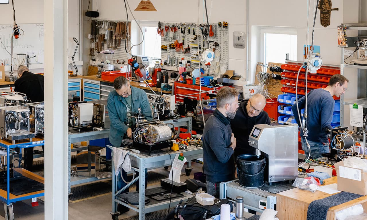 Werkstatt mit Technikern, die Espressomaschinen warten und reparieren; Werkbänke, Werkzeuge und Ersatzteile sichtbar