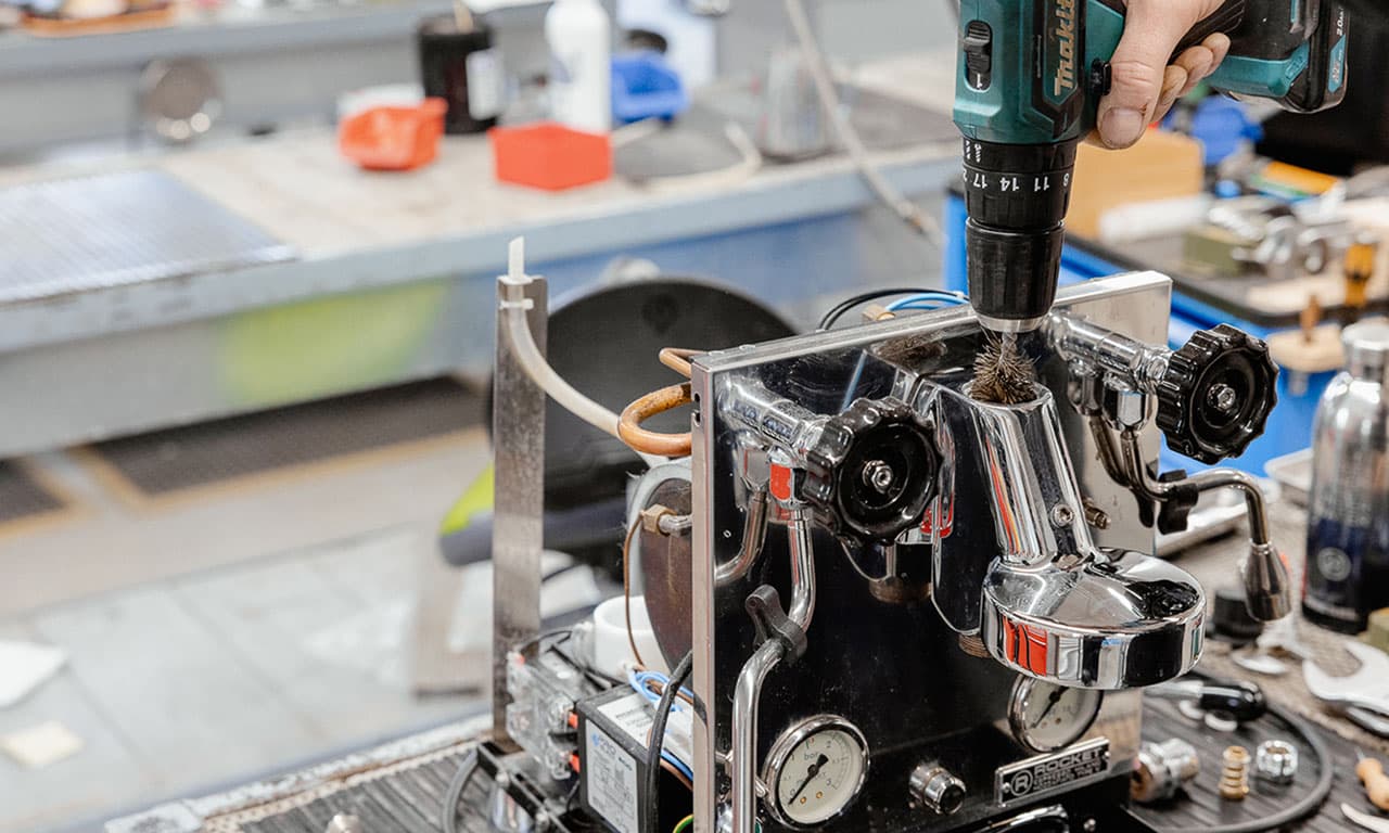 Techniker reinigt und bearbeitet verchromte Espressomaschine mit Bohrmaschine in Werkstatt, Wartung und Service