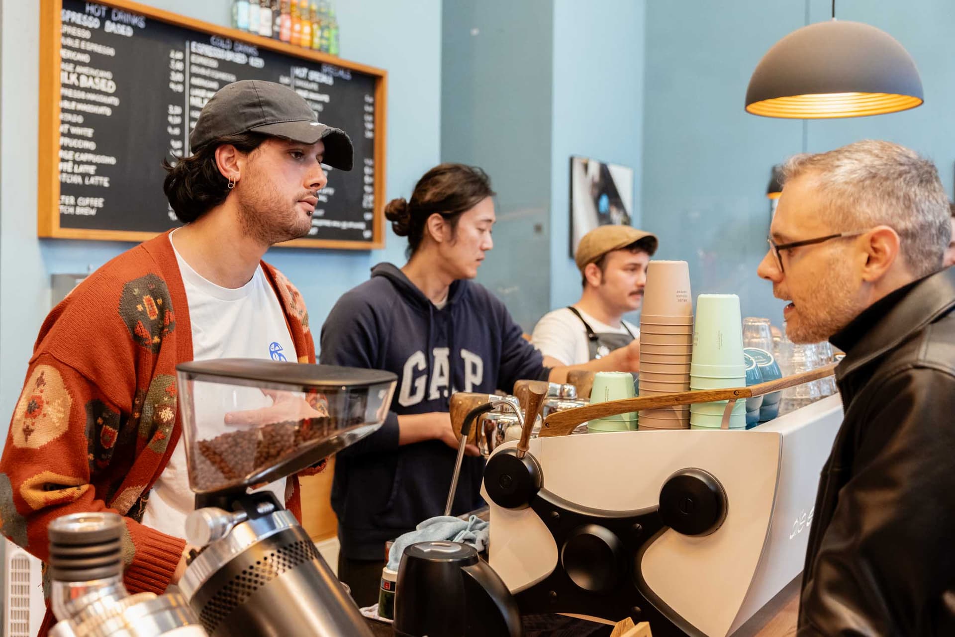 Baristas hinter Espressomaschine bedienen Kundschaft an Theke, Kaffeebohnen, Becherstapel und Menütafel sichtbar
