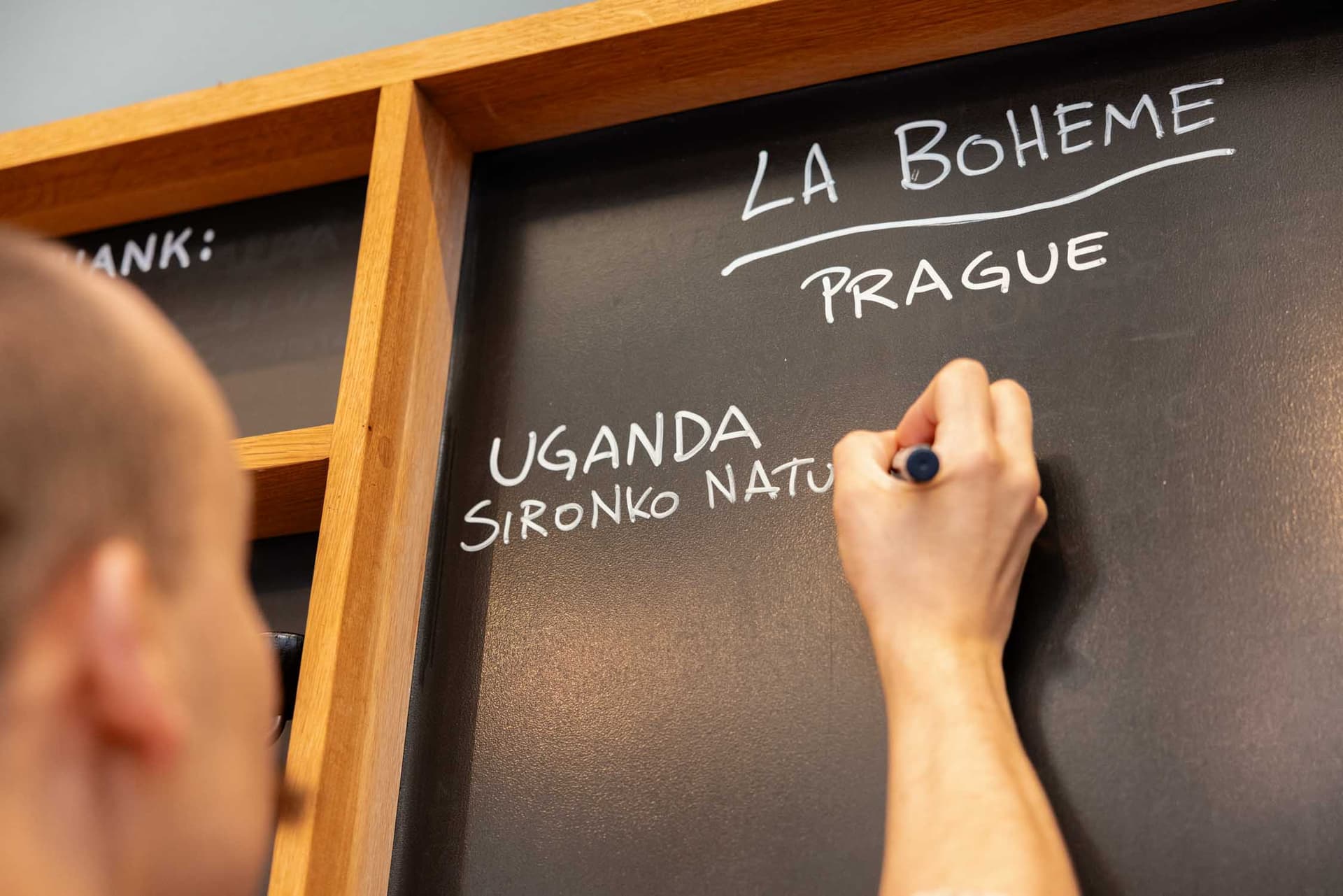 Hand schreibt mit Marker 'La Bohème Prague' auf schwarze Tafel über Holzregal – Bar Takeover Hinweis
