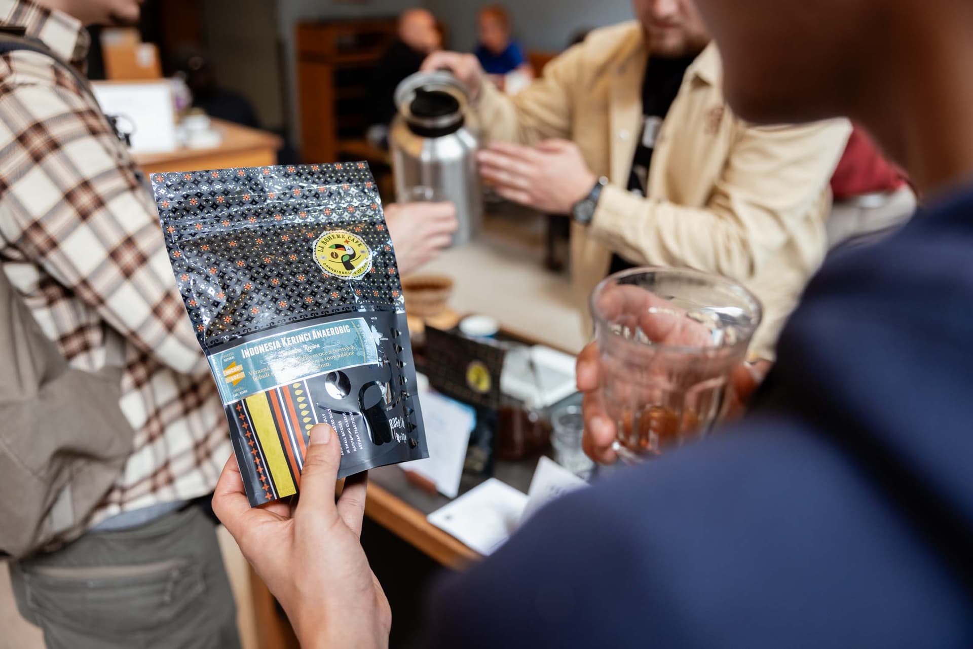 Hand hält verziertes Kaffee-Paket während Barista im Hintergrund Kaffee aus Thermoskanne einschenkt, Bar-Takeover La Bohème