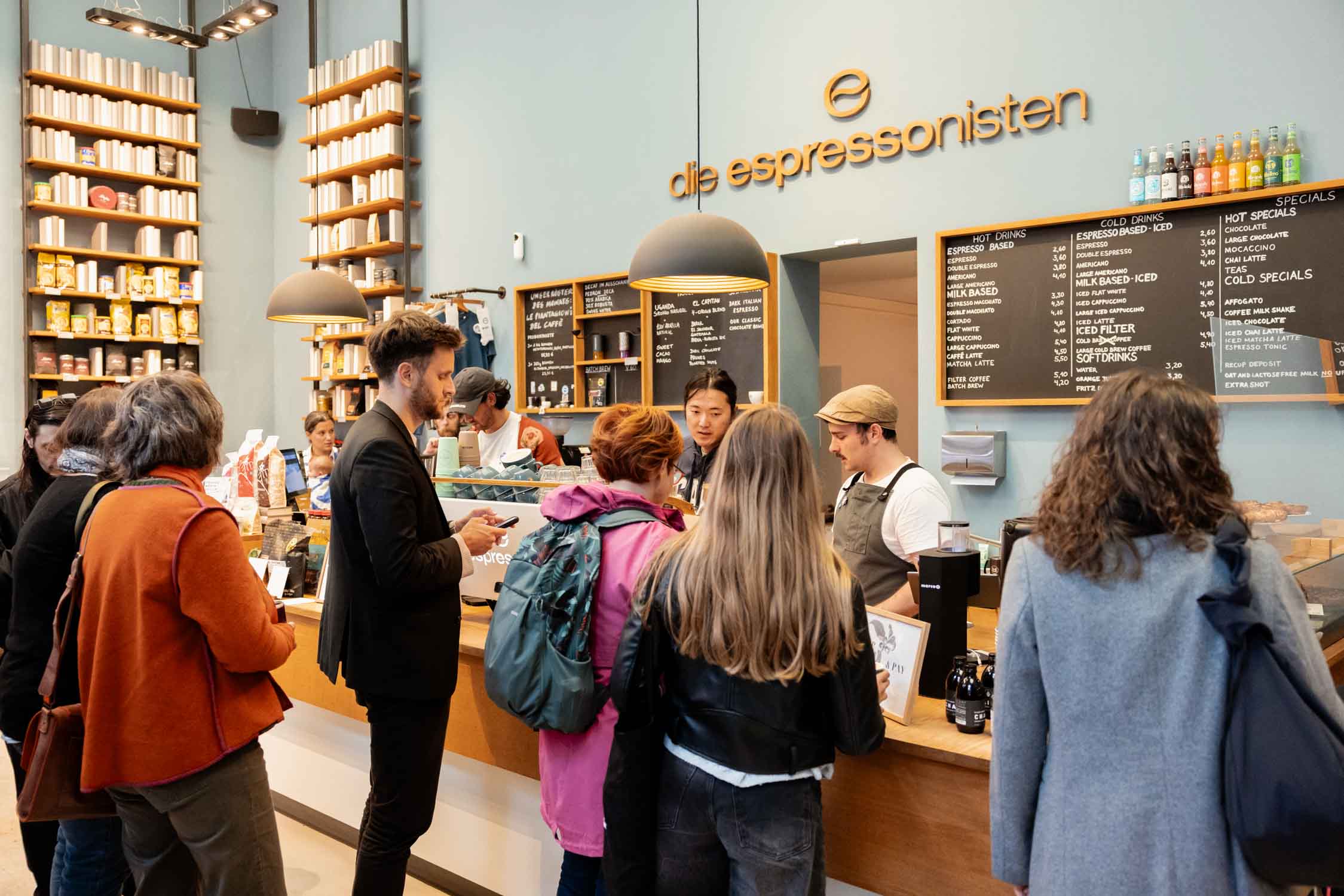 Menschen stehen an der Bar von ‚die espressonisten‘, Barista serviert Kaffee, Regal mit Bohnen und Menütafel